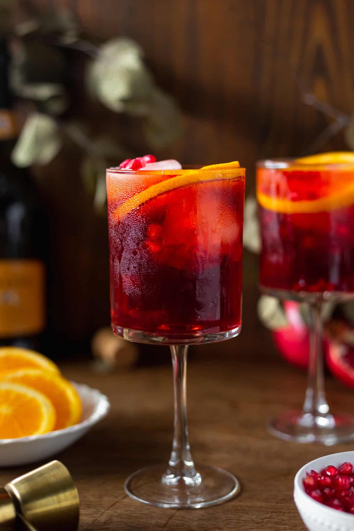 A cocktail glass with pomegranate aperol spritz surrounded by orange and pomegranate seeds