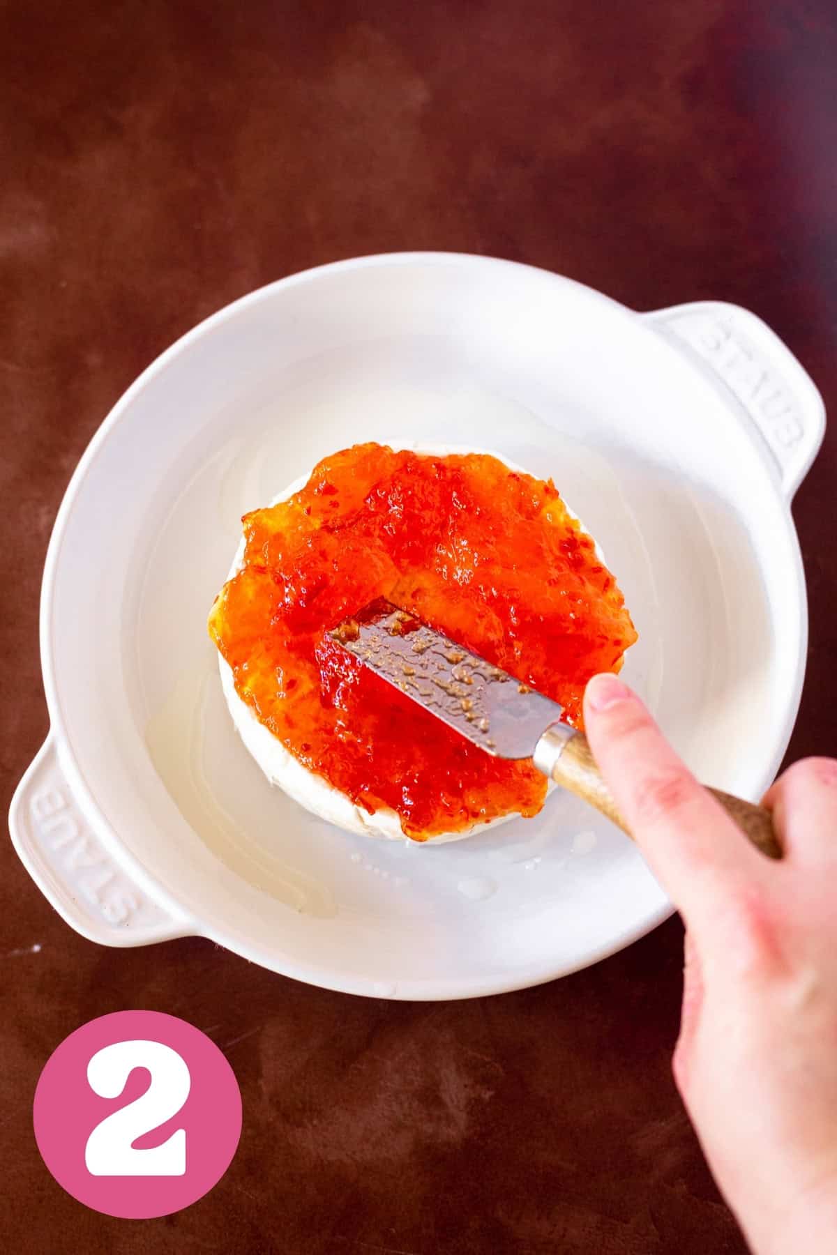 Red pepper jelly being spread on brie with a cheese knife